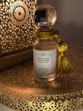 A perfume bottle labeled 'Wrong Side of Customs - Aicha Afterlight' with a gold tassel, set on a decorative gold tray with a warm, patterned background.