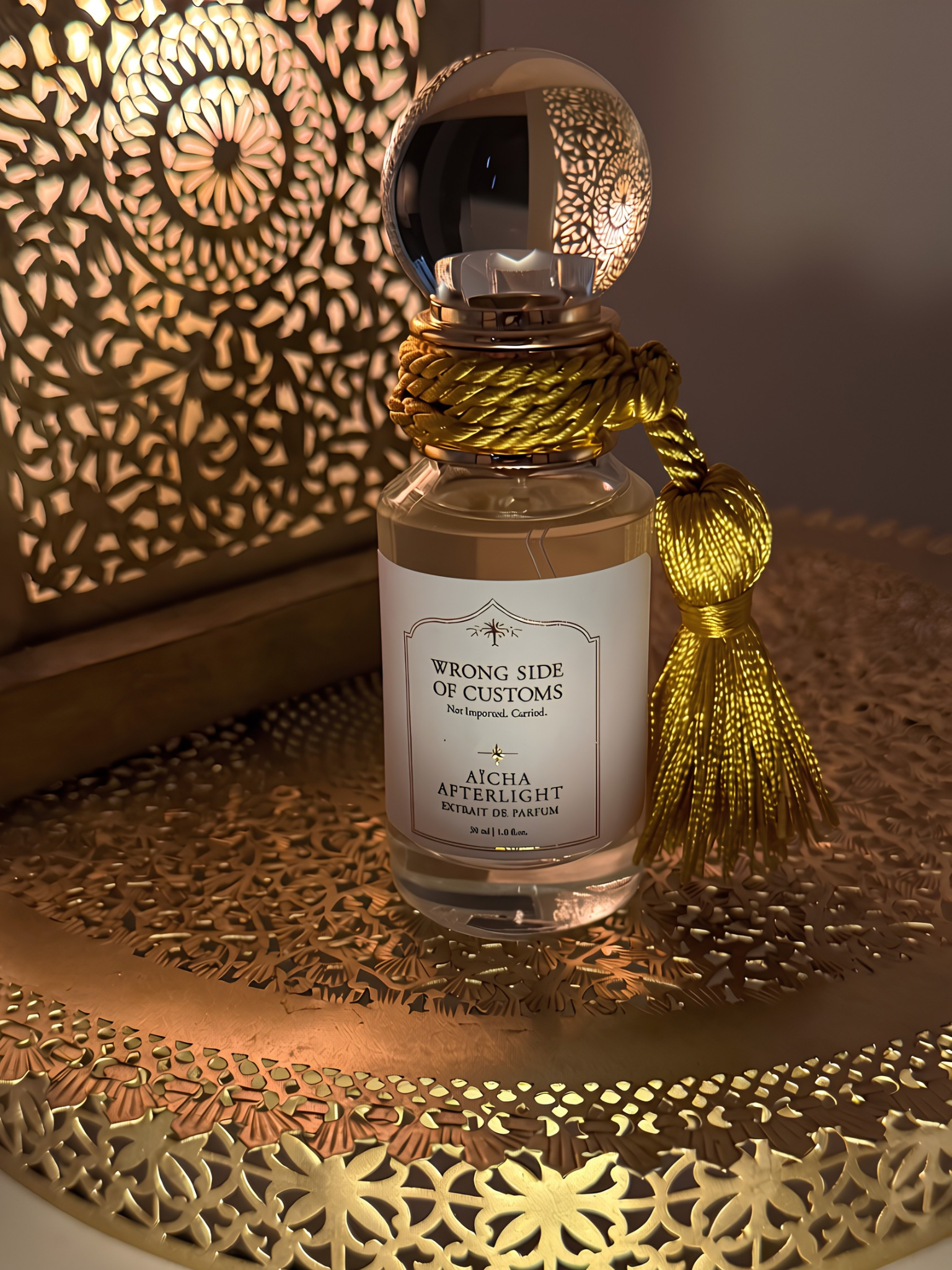 A perfume bottle labeled 'Wrong Side of Customs - Aicha Afterlight' with a gold tassel, set on a decorative gold tray with a warm, patterned background.