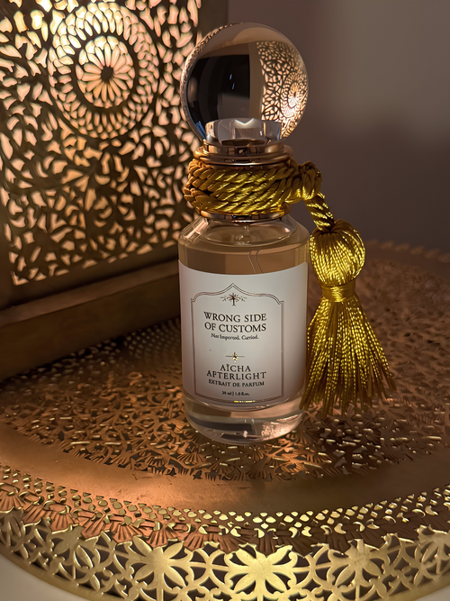 A perfume bottle labeled 'Wrong Side of Customs - Aicha Afterlight' with a gold tassel, set on a decorative gold tray with a warm, patterned background.