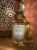 A bottle of Cheri Marrakech 'Wrong Side of Customs' extrait de parfum, featuring a white label and a decorative gold tassel tied around the neck, set against a warm, patterned Moroccan lantern backdrop.