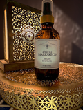 A bottle of Cheri Marrakech Argan Body Oil stands on a decorative gold tray with a warm, ambient background and a hanging lantern casting shadows.