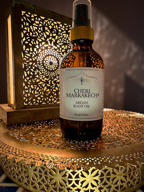 A bottle of Cheri Marrakech Argan Body Oil stands on a decorative gold tray with a warm, ambient background and a hanging lantern casting shadows.