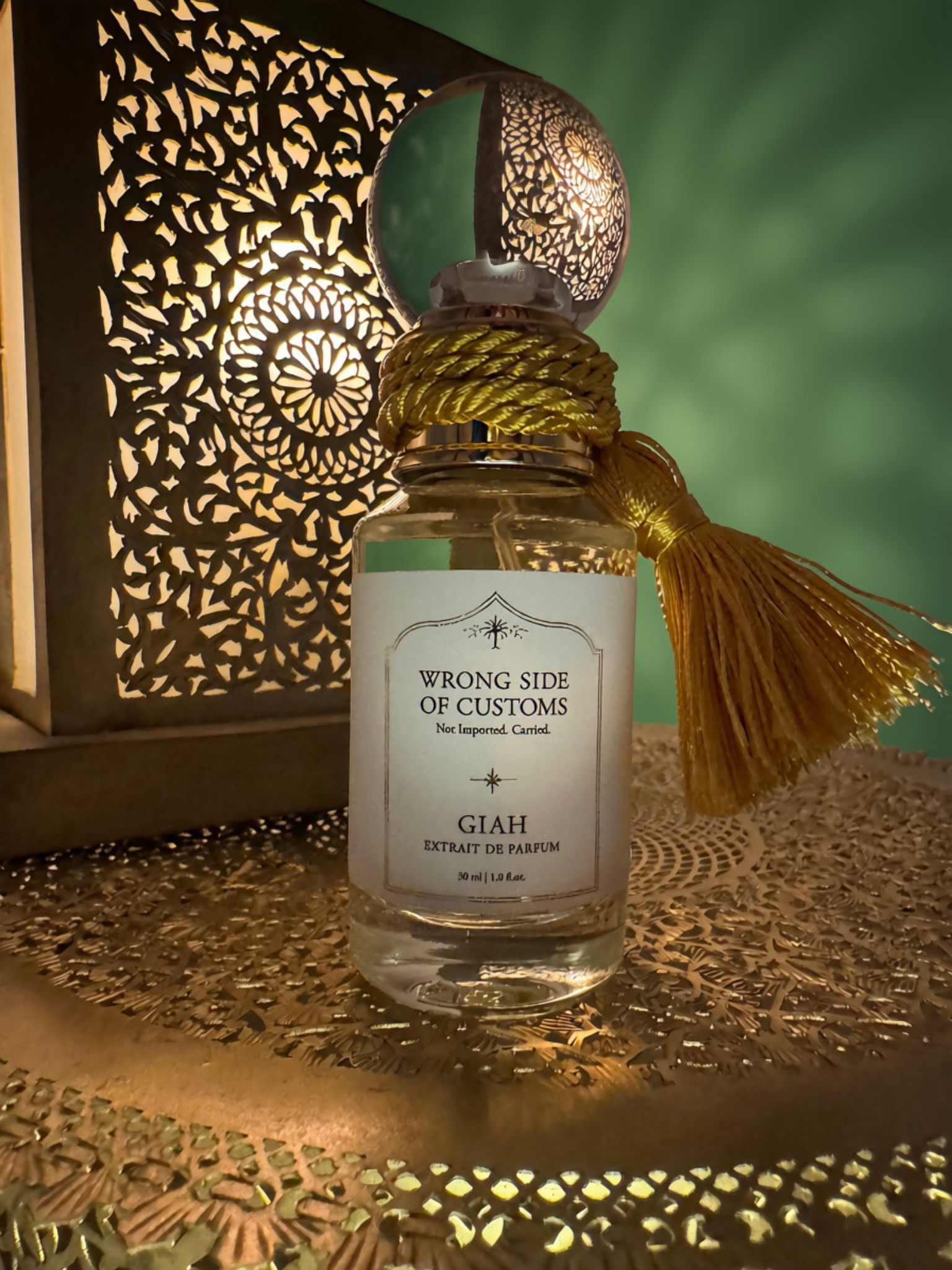 A small glass perfume bottle labeled 'WRONG SIDE OF CUSTOMS' and 'GIAH EXTRAIT DE PARFUM' with a gold tassel and metallic cap, styled on a patterned gold surface beside a glowing cutout lantern against a green backdrop.