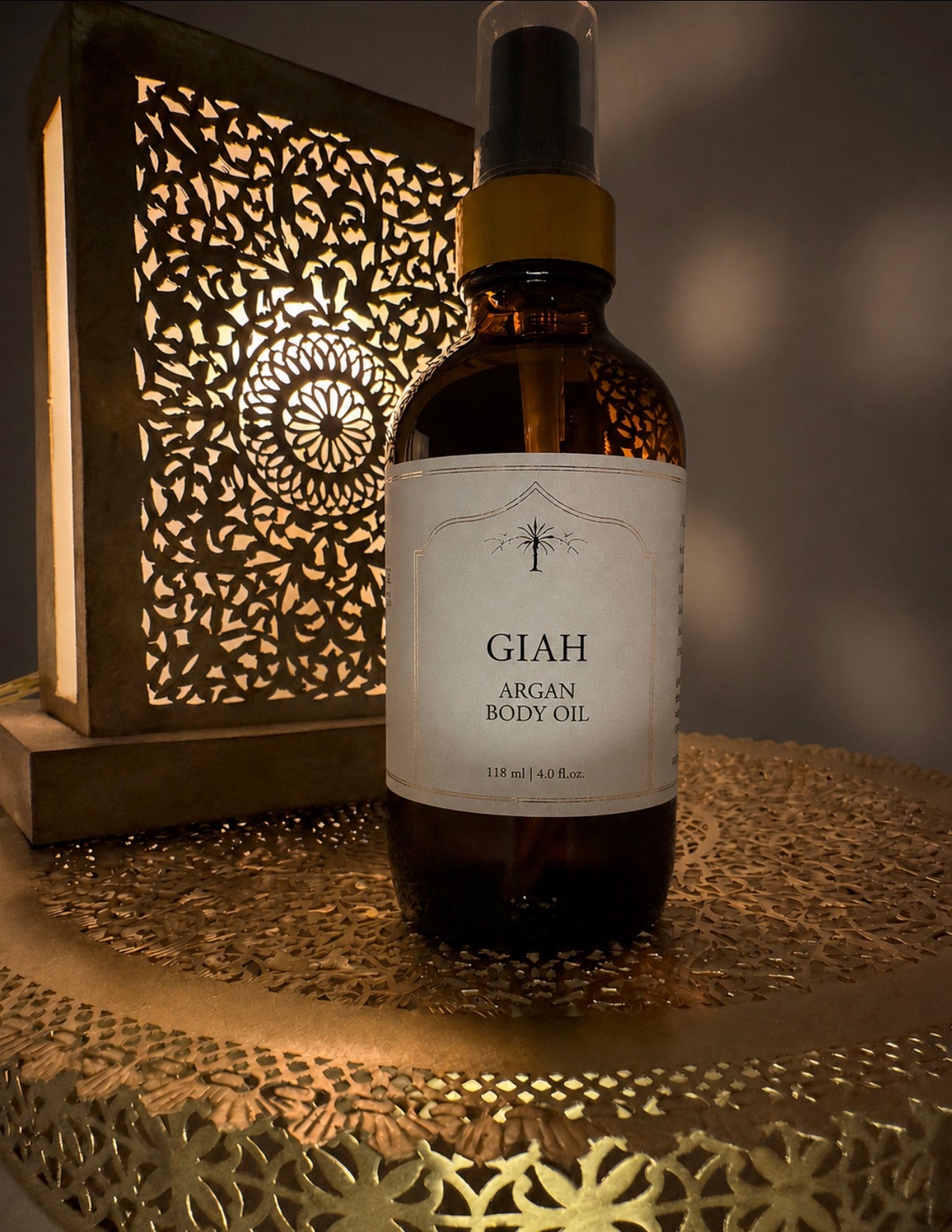 A bottle of GIAH Argan Body Oil stands on a decorative brass tray with a warm, glowing cutout lantern in the background.