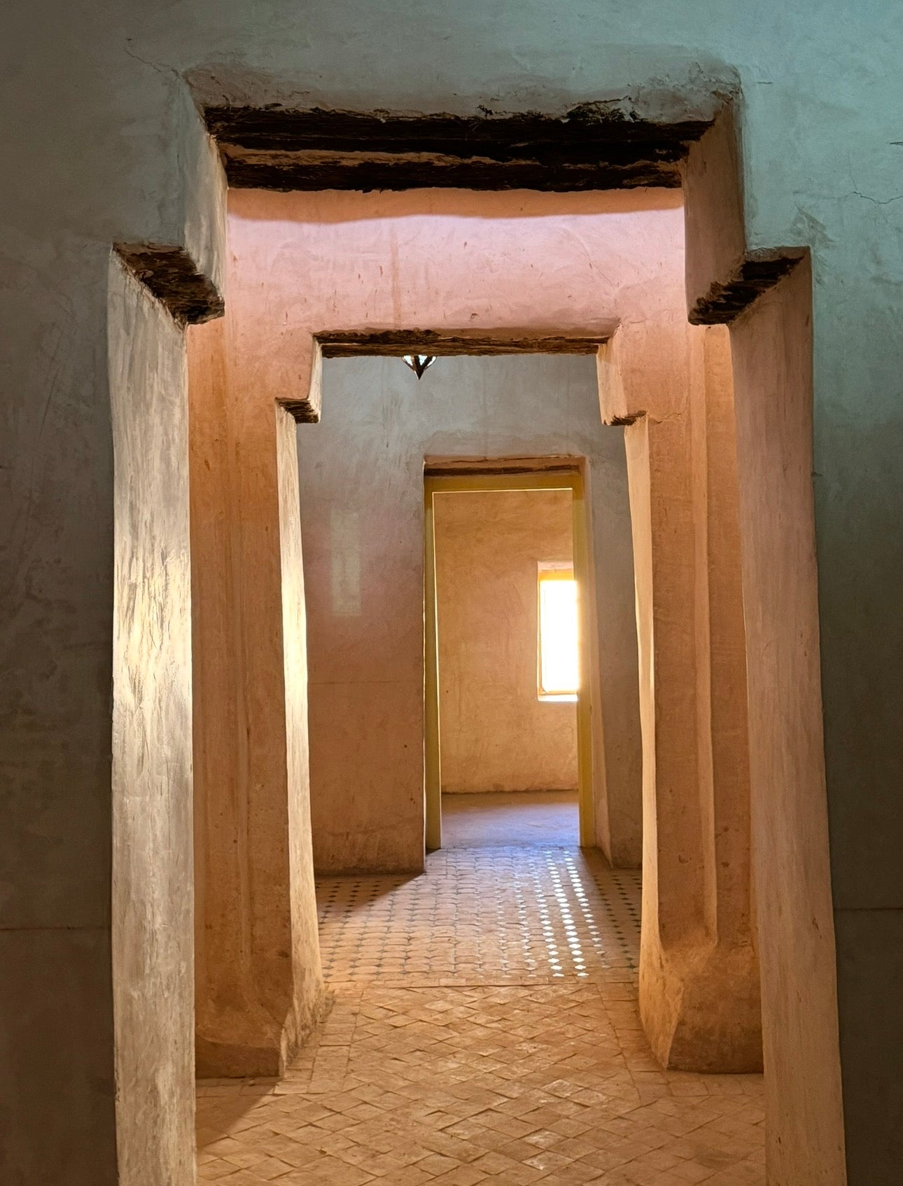 A view looking down a narrow hallway with rough beige stone or plaster walls and pillars, framed by large doorways, with a bright window at the end of the corridor.