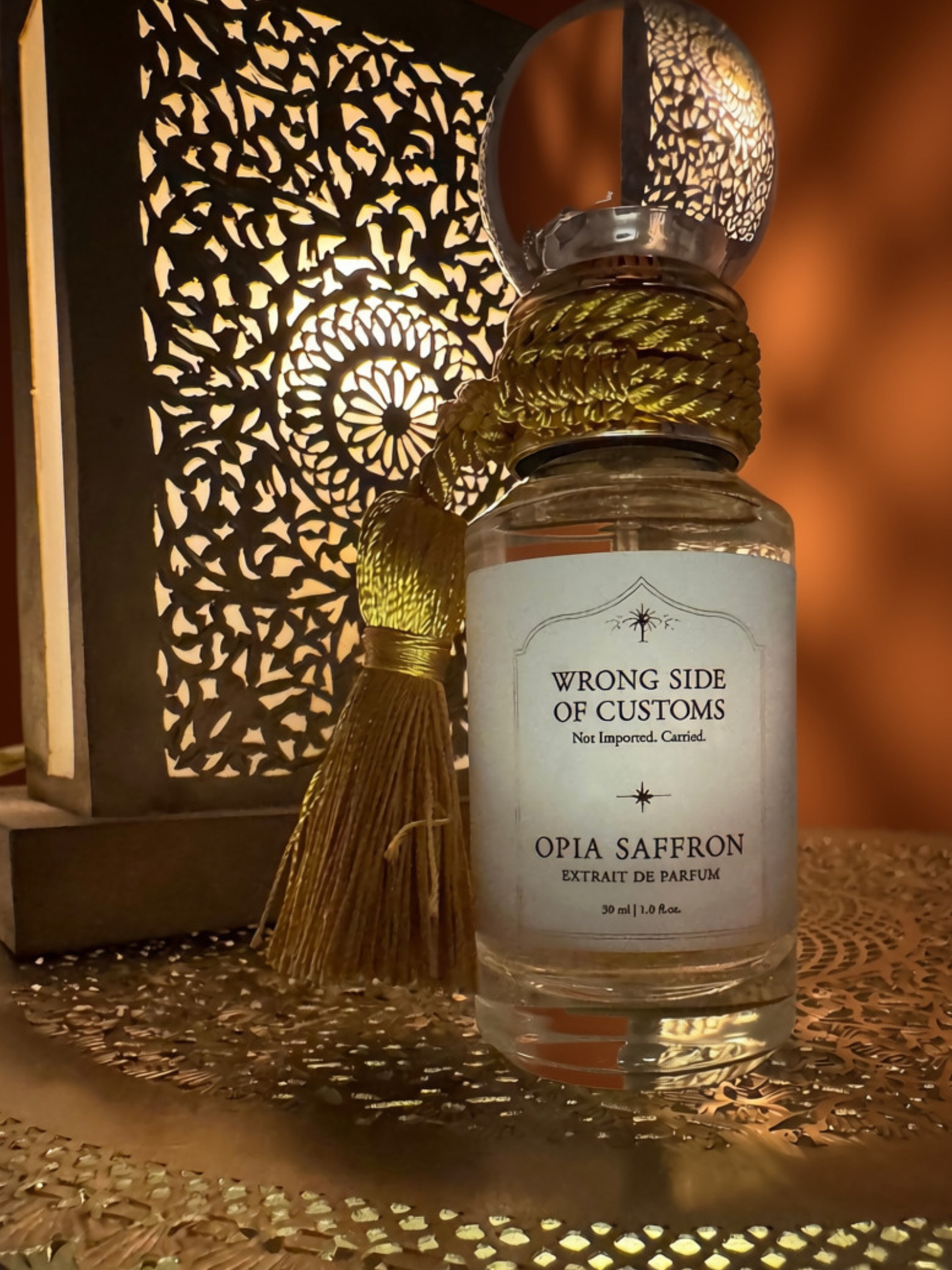 A clear glass perfume bottle labeled 'WRONG SIDE OF CUSTOMS' and 'OPIA SAFFRON' with a white label, wrapped neck with gold cord, topped with a clear ornate cap and a gold tassel, set on a decorative tray beside a glowing cutout lantern.