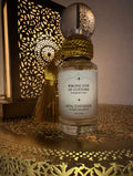 A clear glass perfume bottle labeled 'WRONG SIDE OF CUSTOMS' and 'OPIA STATESIDE' with a white label, wrapped in gold cord, and topped with a spherical cap, set on a decorative gold tray beside a lit cutout lantern.