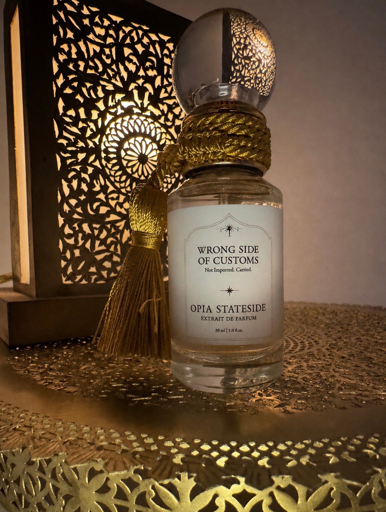 A clear glass perfume bottle labeled 'WRONG SIDE OF CUSTOMS' and 'OPIA STATESIDE' with a white label, wrapped in gold cord, and topped with a spherical cap, set on a decorative gold tray beside a lit cutout lantern.