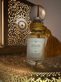 A clear glass perfume bottle with a white label reading 'WRONG SIDE OF CUSTOMS' and 'OPIA OF FEZ', filled with pale golden liquid. The bottle is wrapped with a thick gold cord and has a large gold tassel attached.