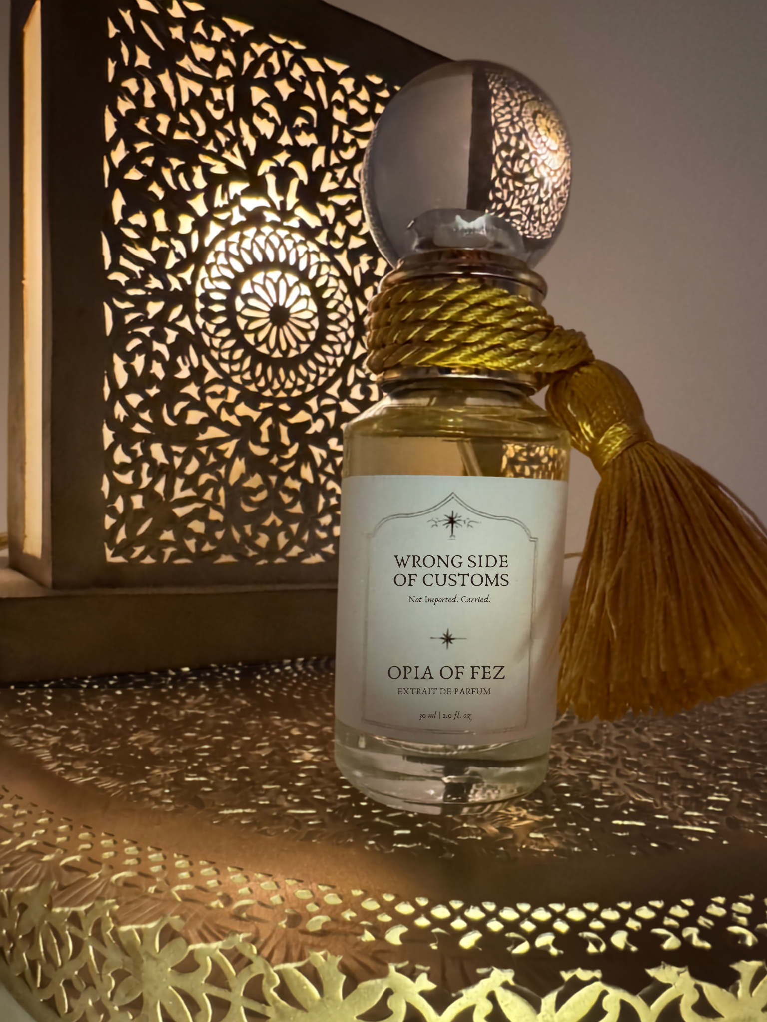 A clear glass perfume bottle with a white label reading 'WRONG SIDE OF CUSTOMS' and 'OPIA OF FEZ', filled with pale golden liquid. The bottle is wrapped with a thick gold cord and has a large gold tassel attached.