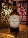 A bottle of Souk Lil Argan Body Oil stands on a metallic surface with a decorative Moroccan-style lantern and patterned tile background.