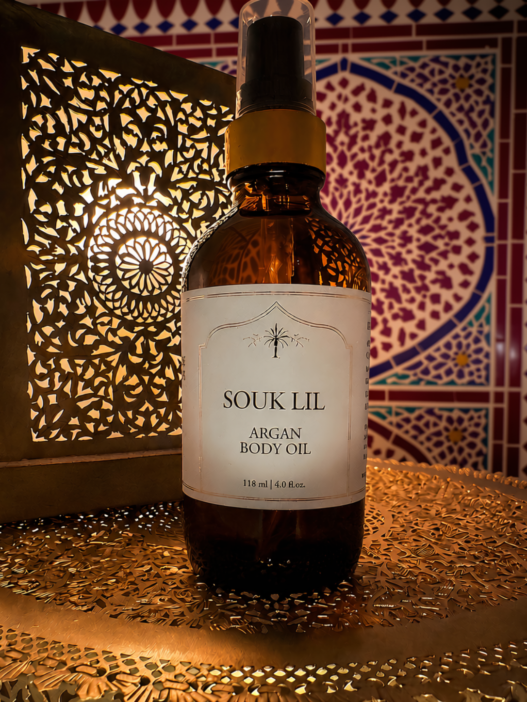 A bottle of Souk Lil Argan Body Oil stands on a metallic surface with a decorative Moroccan-style lantern and patterned tile background.