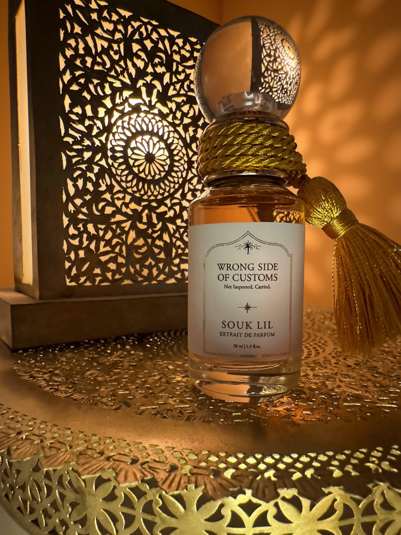 A clear glass perfume bottle labeled 'WRONG SIDE OF CUSTOMS SOUK LIL' sits on a decorative gold-toned tray with a matching tassel and a warm glowing cutout lantern in the background.