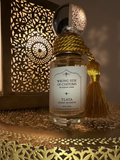A bottle of TLATA Wrong Side of Customs Extrait de Parfum with a white label, gold rope stopper, and a gold tassel, set against a warm, illuminated Moroccan-style lantern background.