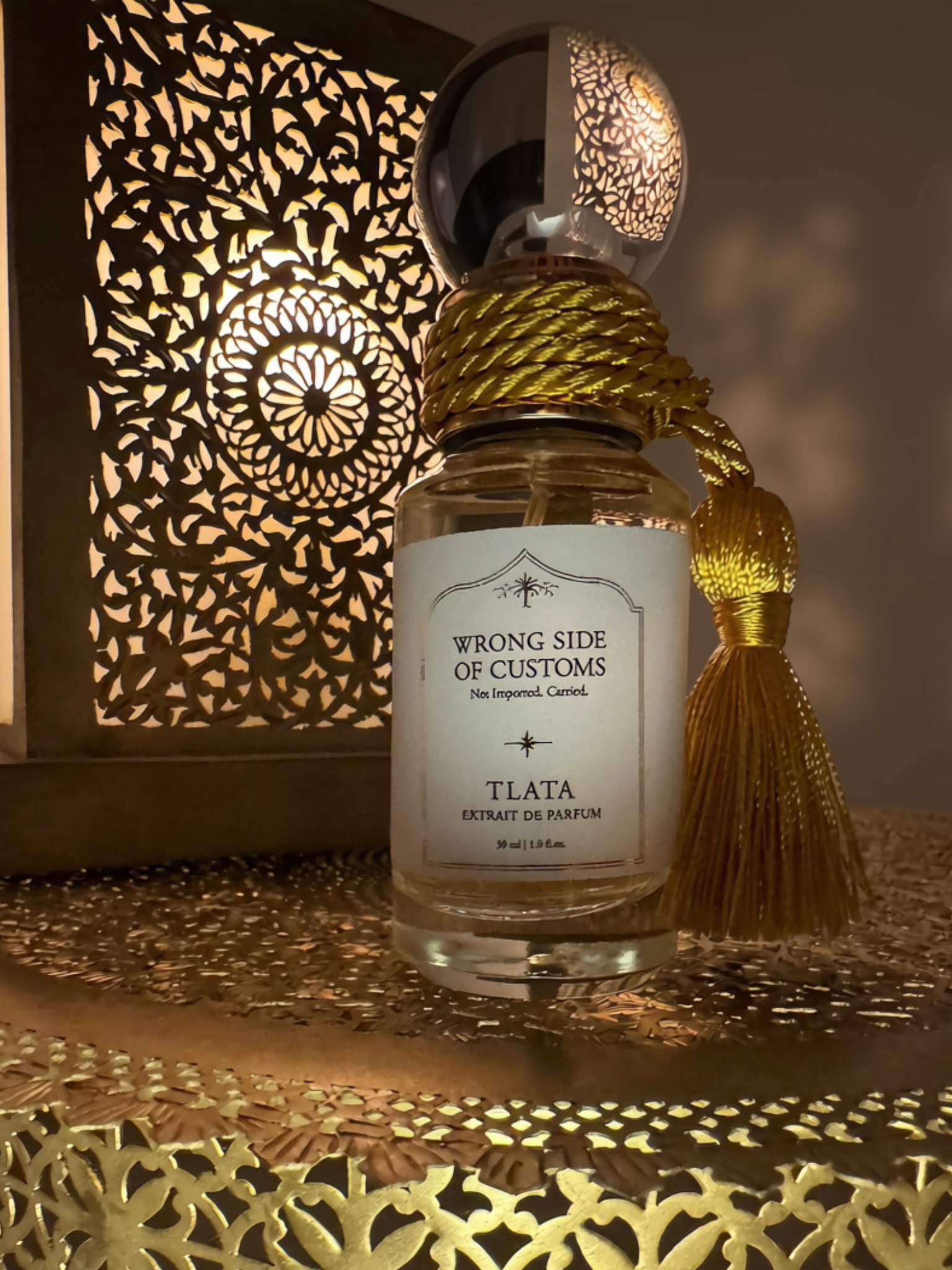 A bottle of TLATA Wrong Side of Customs Extrait de Parfum with a white label, gold rope stopper, and a gold tassel, set against a warm, illuminated Moroccan-style lantern background.